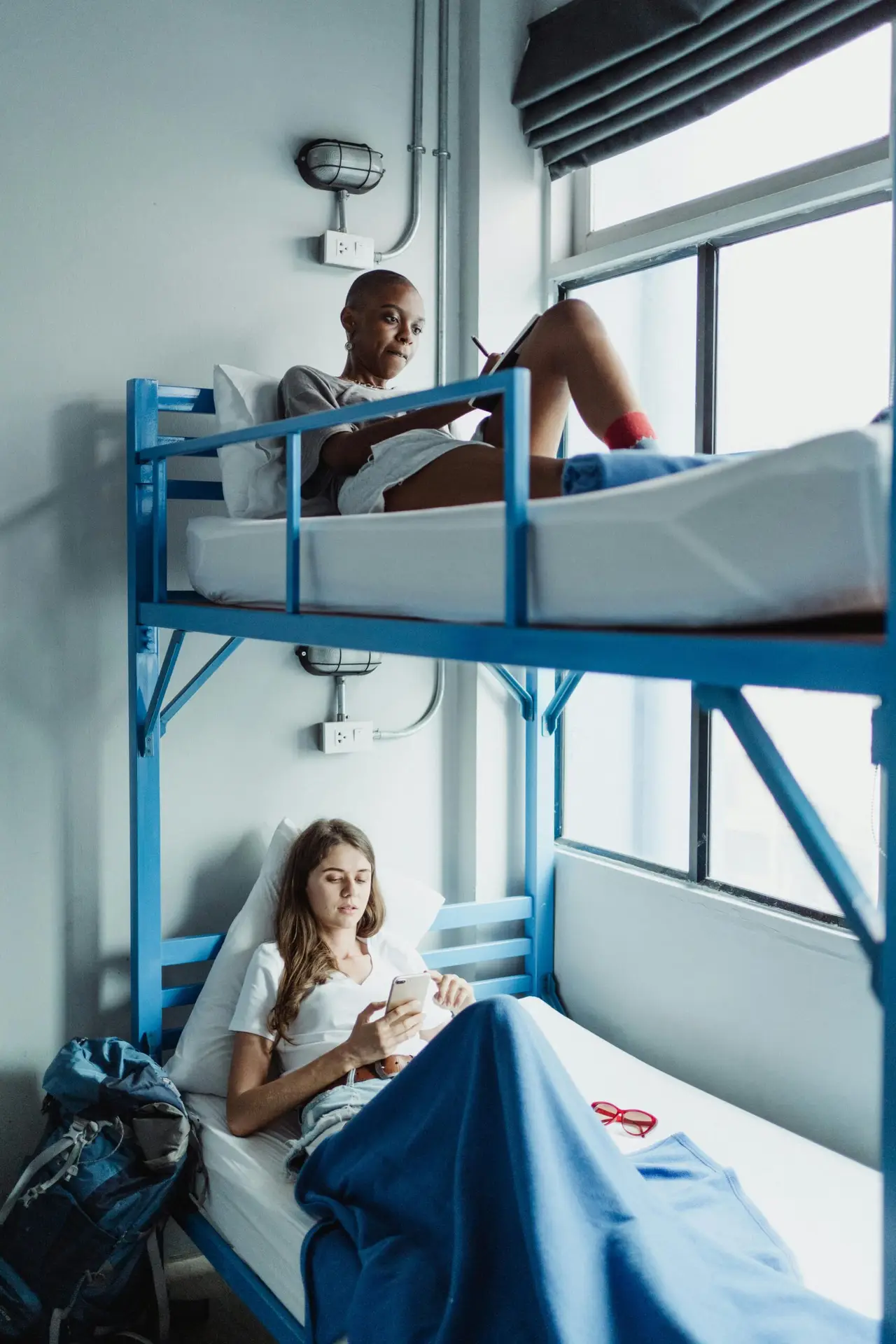 Two women relaxing in bright hostel room bunk beds with backpacks and natural light.